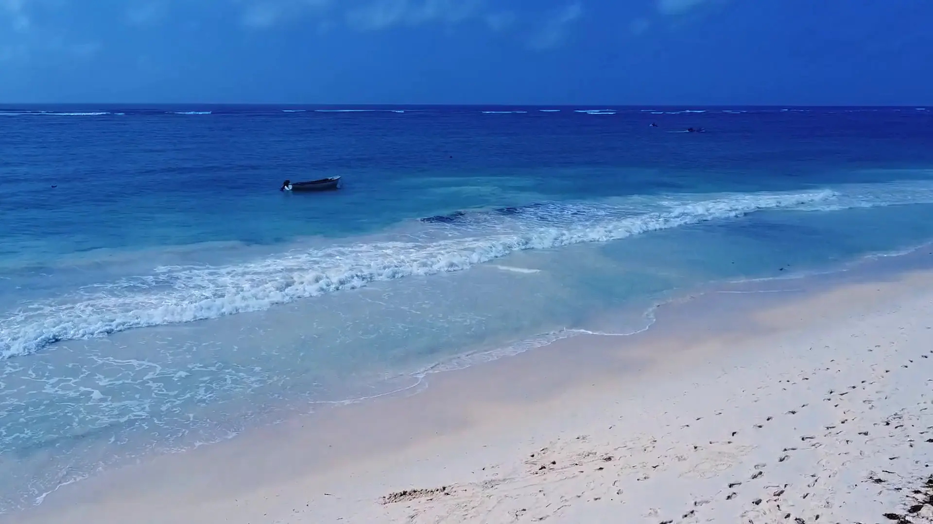 Slow low aerial along white sand as turquoise waves roll in over the shallows, a small boat moored beyond and the reef line on the horizon.