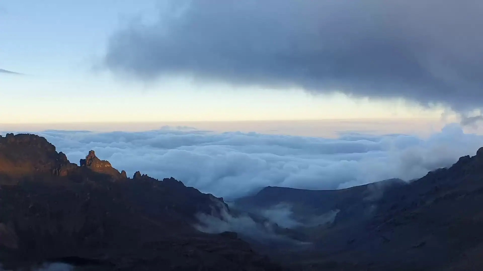 Dawn over a sea of cloud from high on Mount Kenya: early light catches jagged rocky peaks, mist drifts through the valley below and a small blue tent sits on the stony ground.