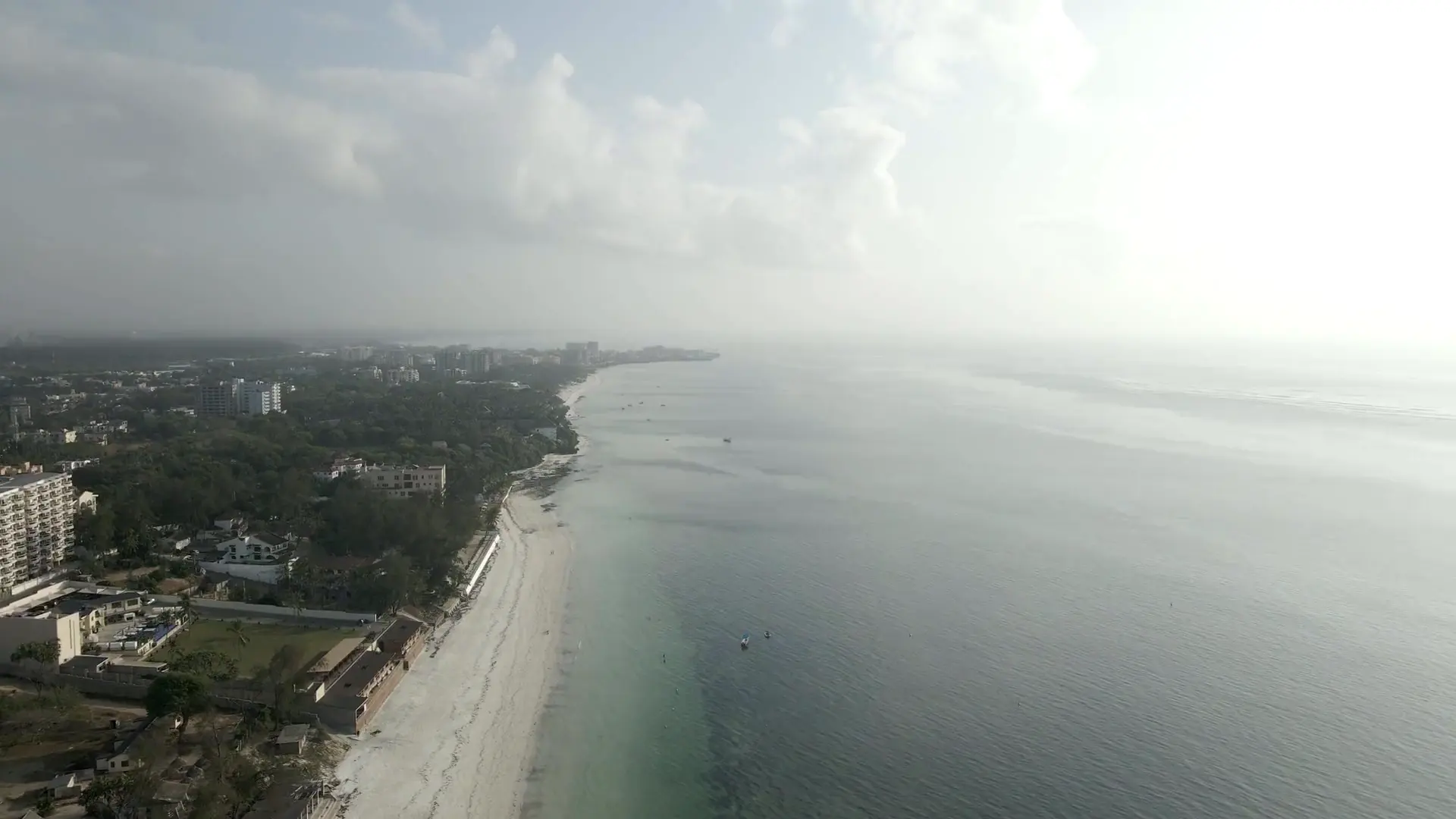 Hazy aerial along the Nyali shoreline in Mombasa: a thin white beach curving north past apartment blocks and trees beside calm, pale sea.