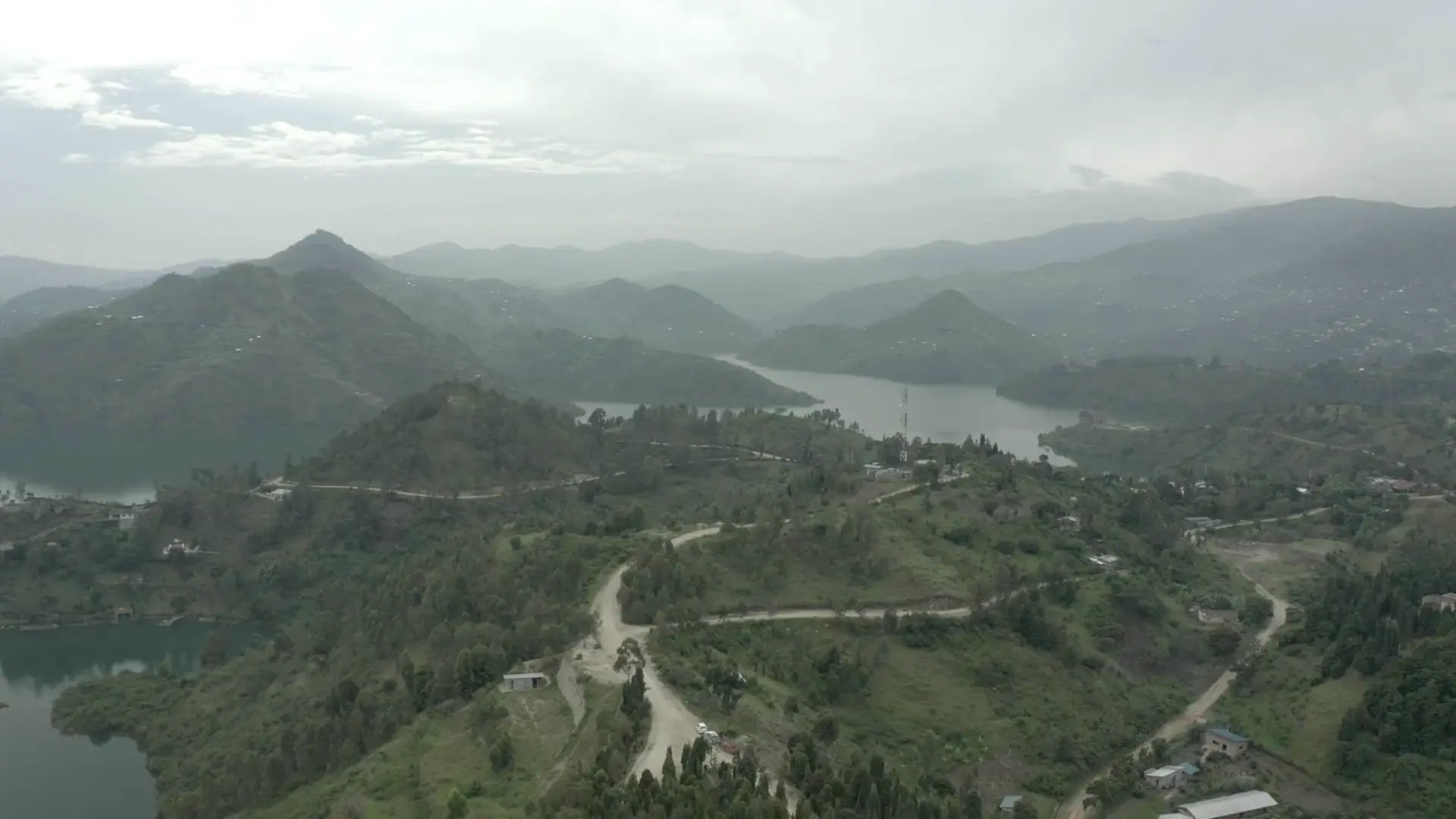 Aerial view over green hills, winding dirt roads and a long lake between steep ridges under soft grey cloud.
