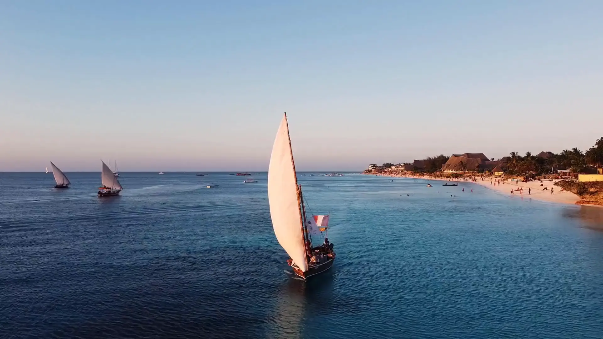 A lateen-sailed dhow glides past a white-sand beach and palm-thatched shore in late afternoon light.
